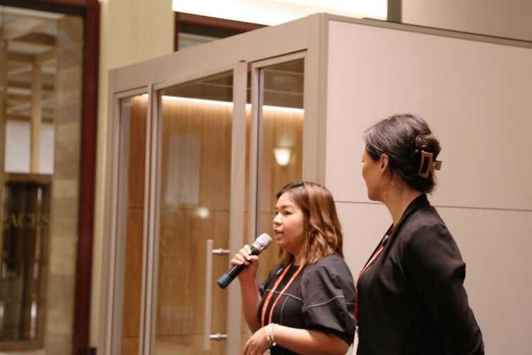Gia Kieu speaking at the NeoCon reveal event, with her winning Zen Garden Pod design displayed behind her.